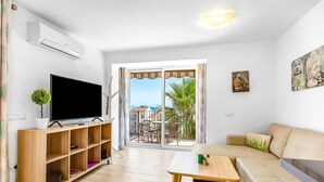 A living room with a TV, wooden shelves, a sofa, a small table, and a balcony with a view of palm trees and the sea. - Holiday Apartment 'Torreblanca Deluxe Holiday Home' with Sea View, Shared Pool & Wi-Fi (Fuengirola)