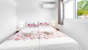 A bedroom with a bed, two bedside tables, an air conditioning unit, a window with curtains, and a view of greenery outside.