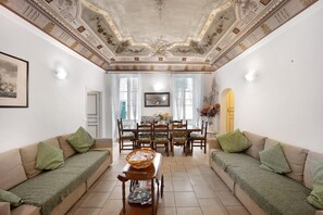 Ein Zimmer mit einer Couch, einem Esstisch und Stühlen, einer Decke mit aufwendigen Verzierungen und einem gerahmten Bild an der Wand.