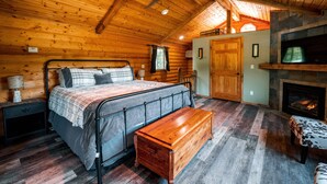 Een slaapkamer in een blokhut met een bed, een bankje, houten wanden, een plafondventilator en een open haard.
