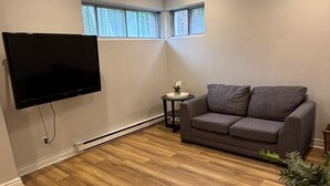 A living room with a flat-screen TV, grey sofa, wooden flooring, and a small side table with a vase.