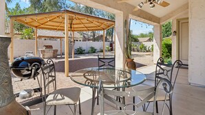 Un patio al aire libre con una mesa y sillas de vidrio, una pérgola de madera, un ventilador de techo y una parrilla.