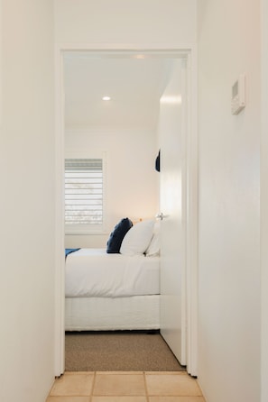 A narrow hallway leading to a bedroom with a bed, a window with blinds, and a thermostat on the wall. - Commodore 7 80 Noosa Parade (Noosa Heads)