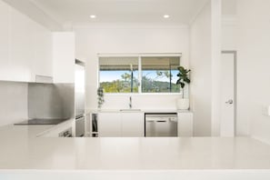 Una cocina moderna con armarios blancos, una campana extractora de acero inoxidable, lavavajillas y una ventana con vista a zonas verdes.