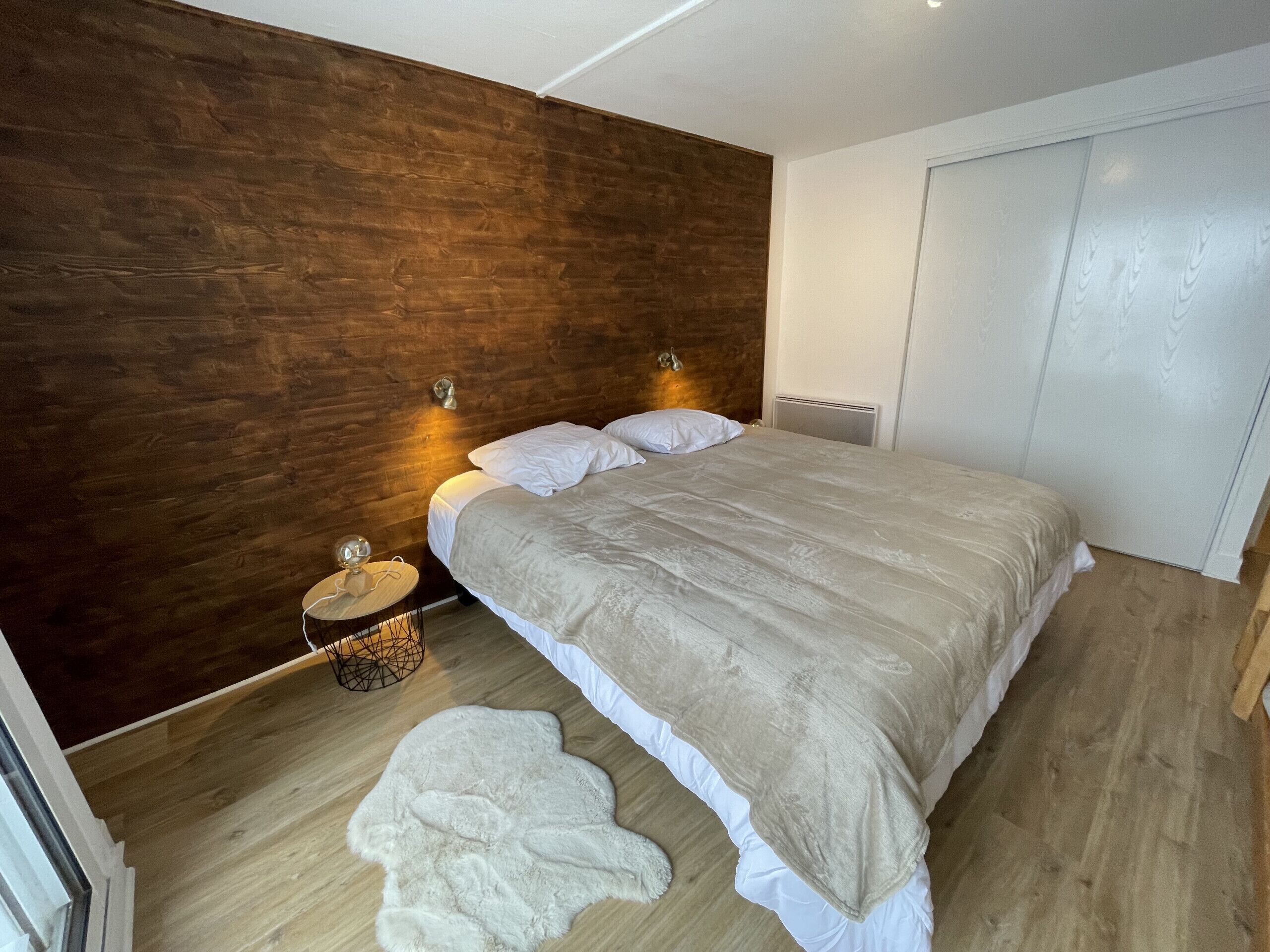 Un dormitorio con cabecera de madera, cama con ropa de cama blanca, una mesita redonda con una lámpara y una alfombra blanca en el suelo.