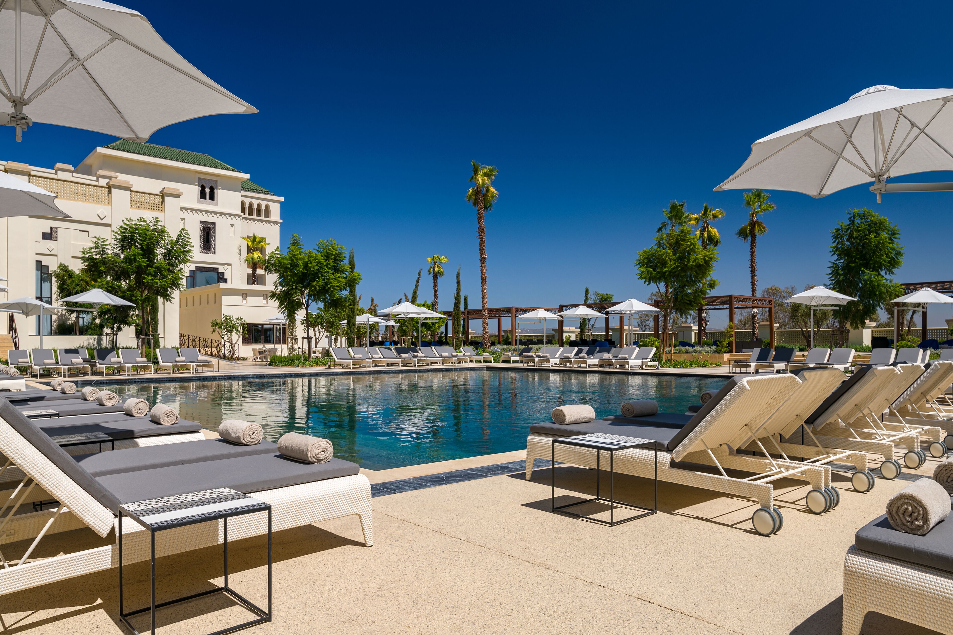 A poolside area with lounge chairs and umbrellas, a clear blue sky, and palm trees.