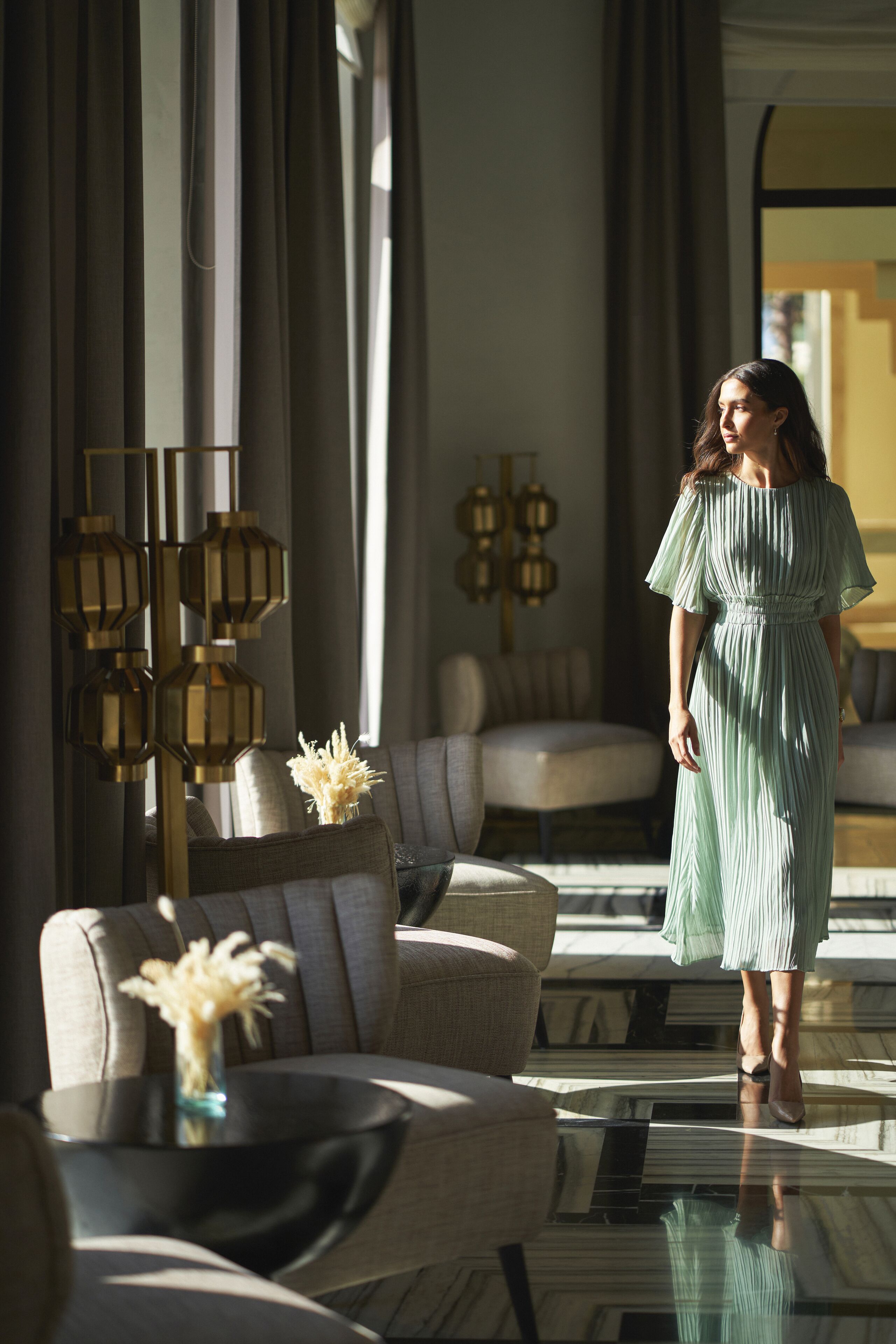 A woman in a green dress standing in a modern living room with a sofa, a glass coffee table, and decorative lanterns.