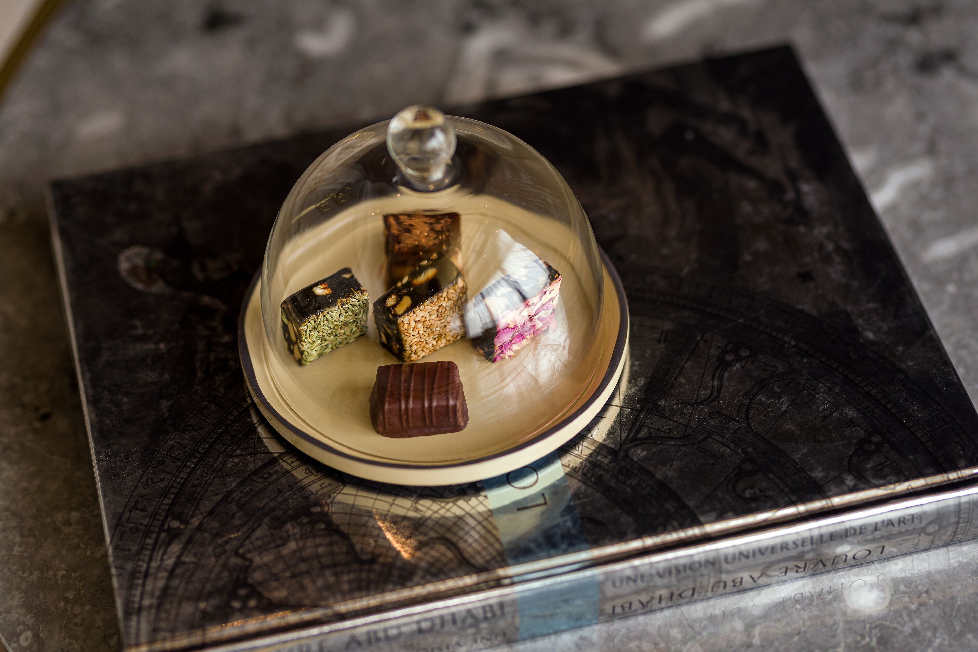 A dessert display with various sweets under a glass dome, placed on a book with visible text.