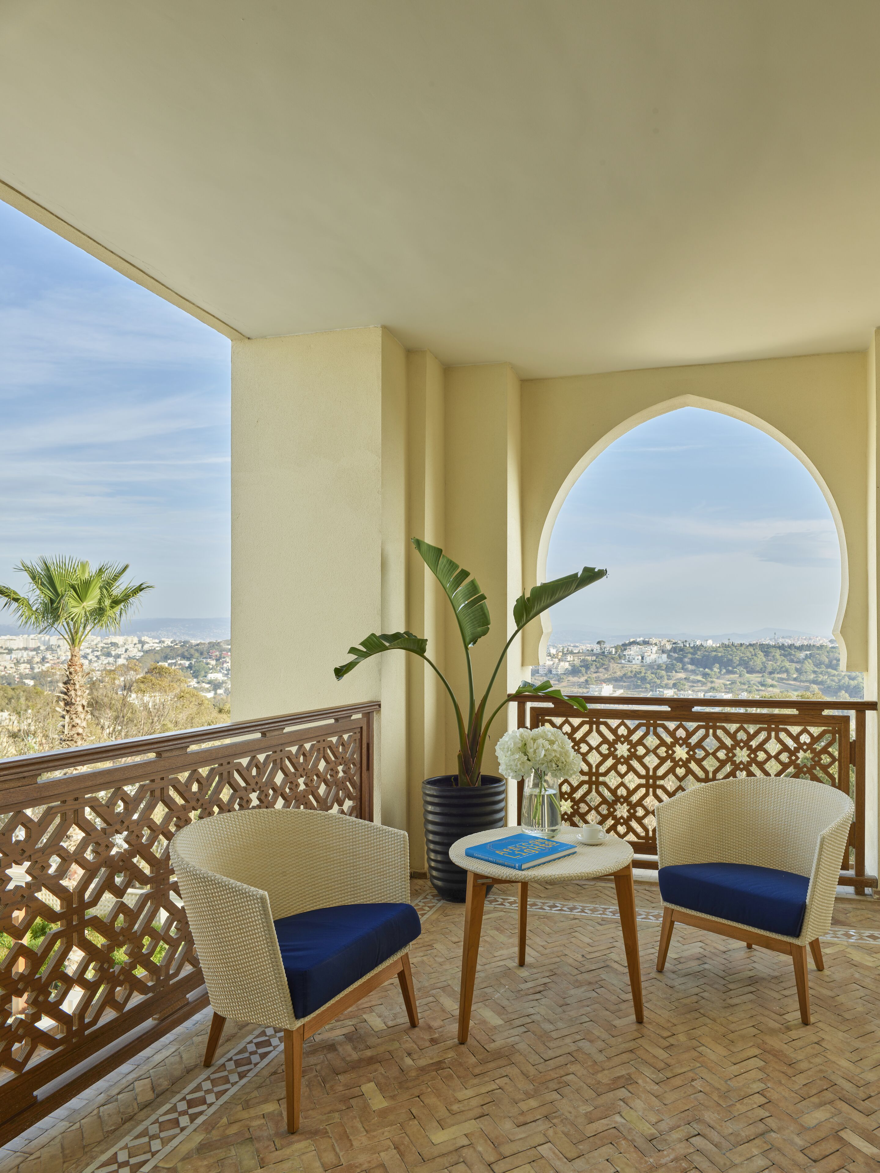 Un balcon avec deux fauteuils en osier, une petite table sur laquelle se trouvent un livre et un vase de fleurs, et une vue sur le paysage urbain.
