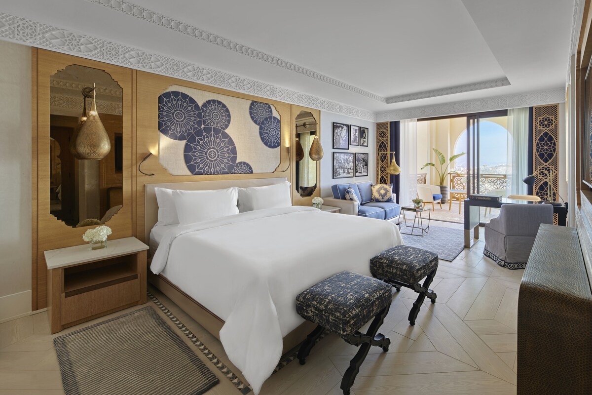 Een hotelkamer met een groot bed, twee fauteuils, een bureau met stoel, een kleine tafel met vaas, en een balkon met uitzicht.