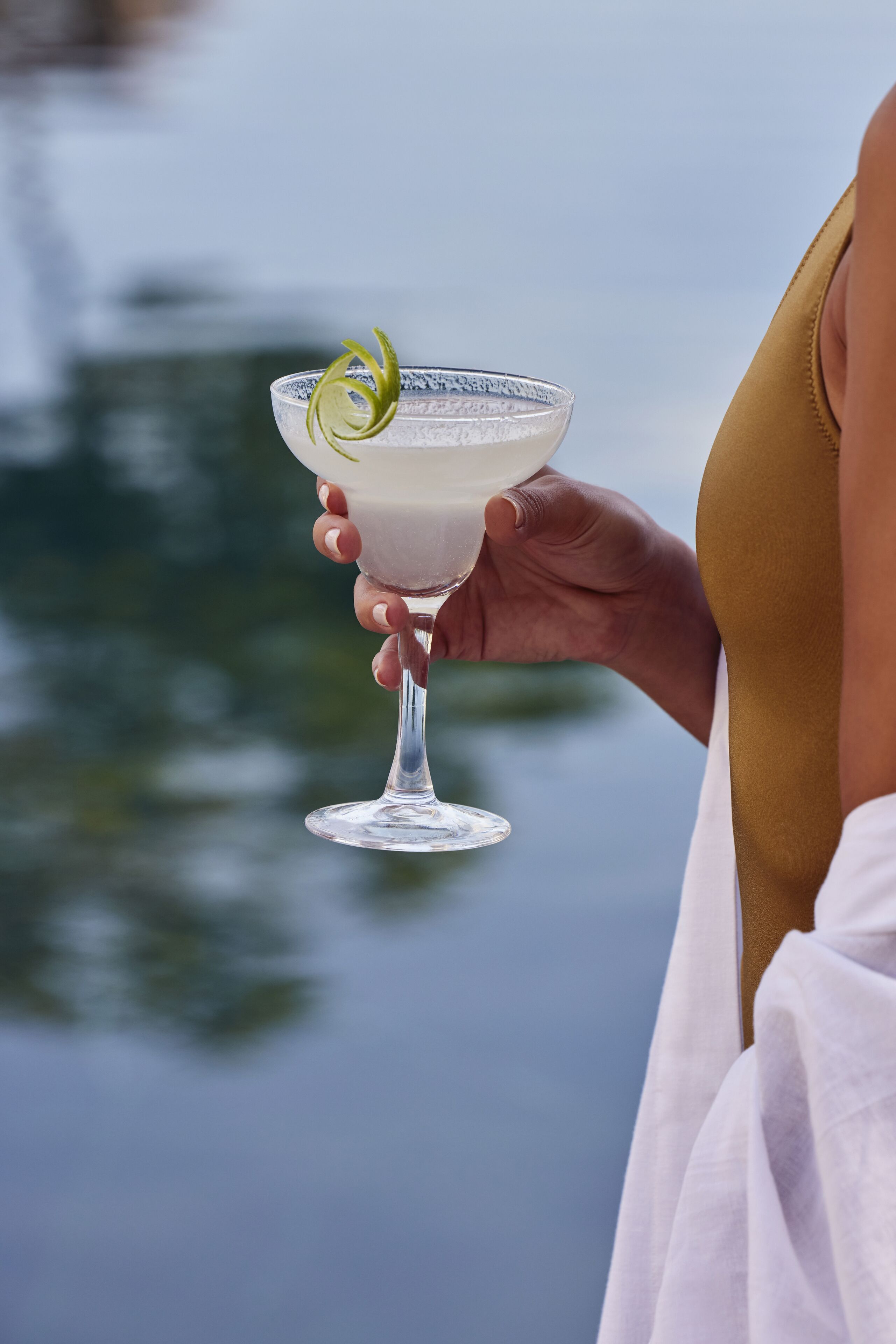A person holding a cocktail glass with a garnish, with a blurred outdoor background.