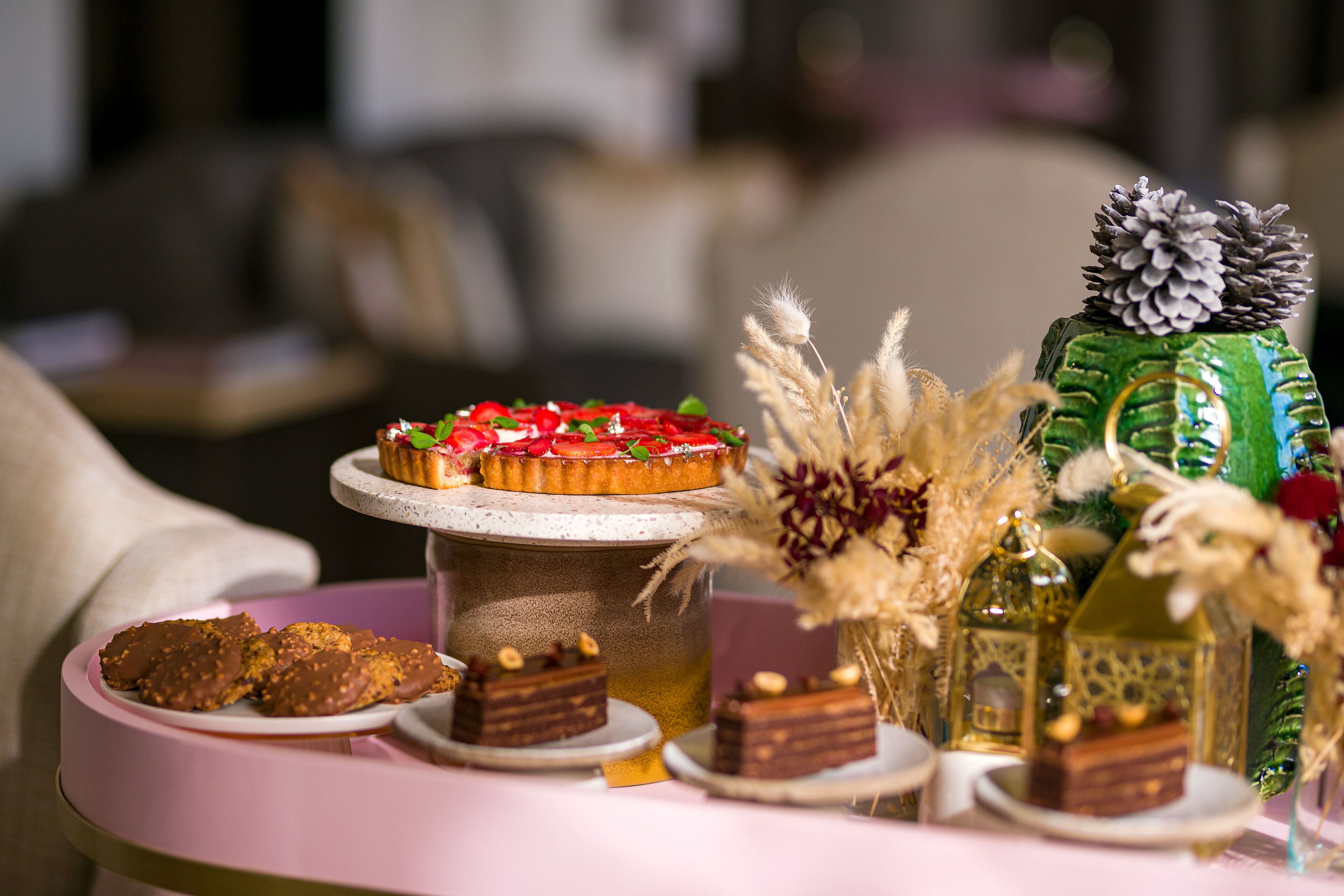 Un tavolo con dolci, torte e pasticcini assortiti, un vaso decorativo con piante essiccate e una bottiglia verde con un pigna sopra.