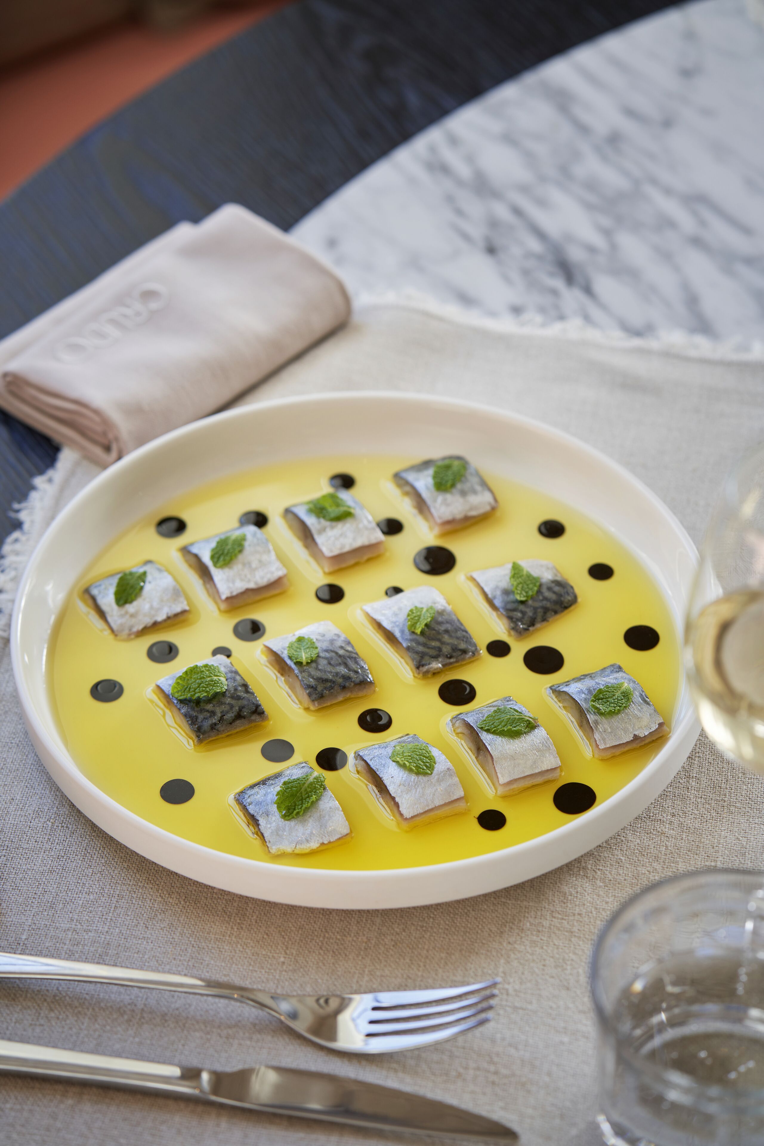 A plate of food with small fish pieces and mint leaves, accompanied by a fork and a glass of white wine.
