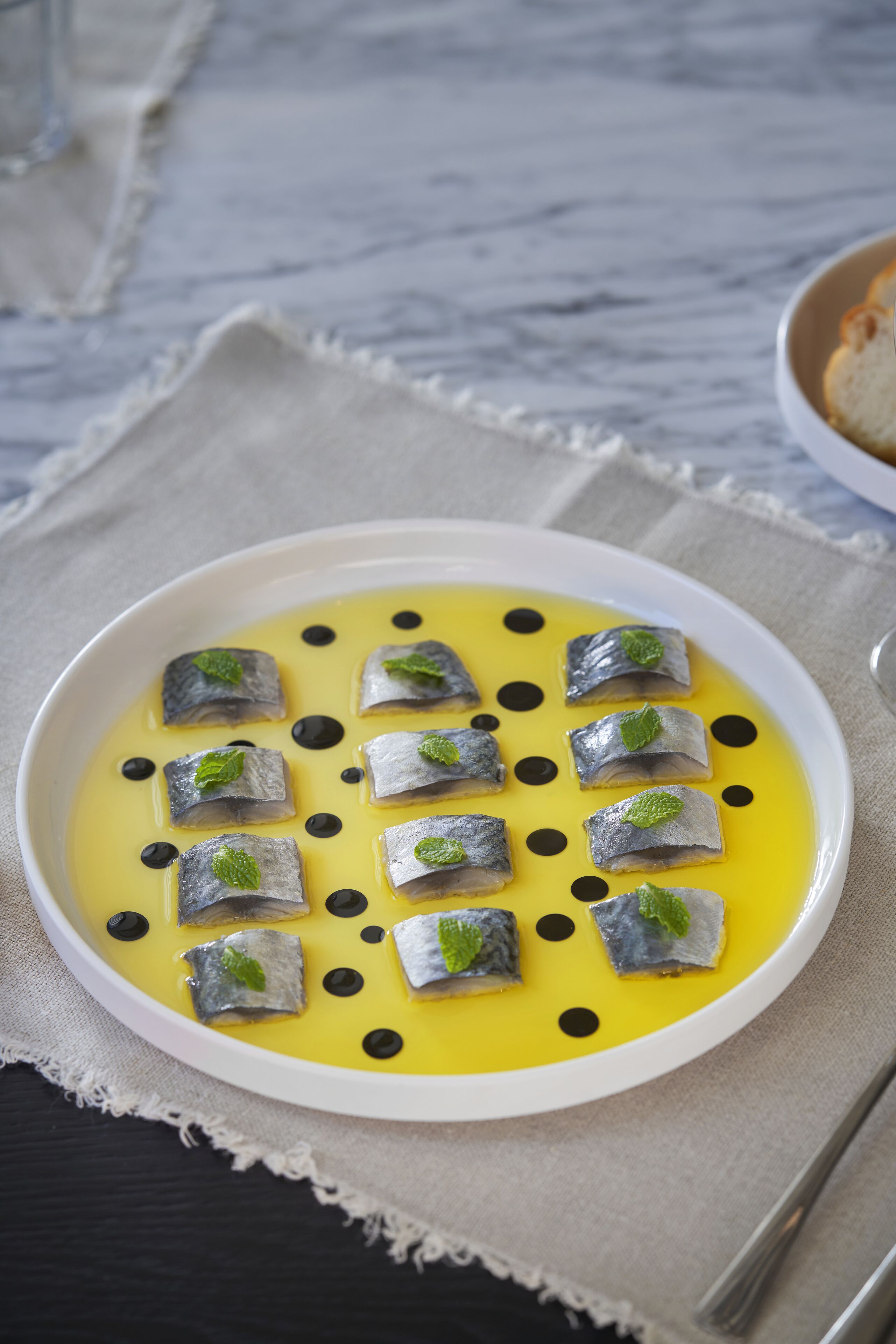 A white plate with small fish garnished with mint leaves, served on a yellow sauce with black dots, on a beige cloth.