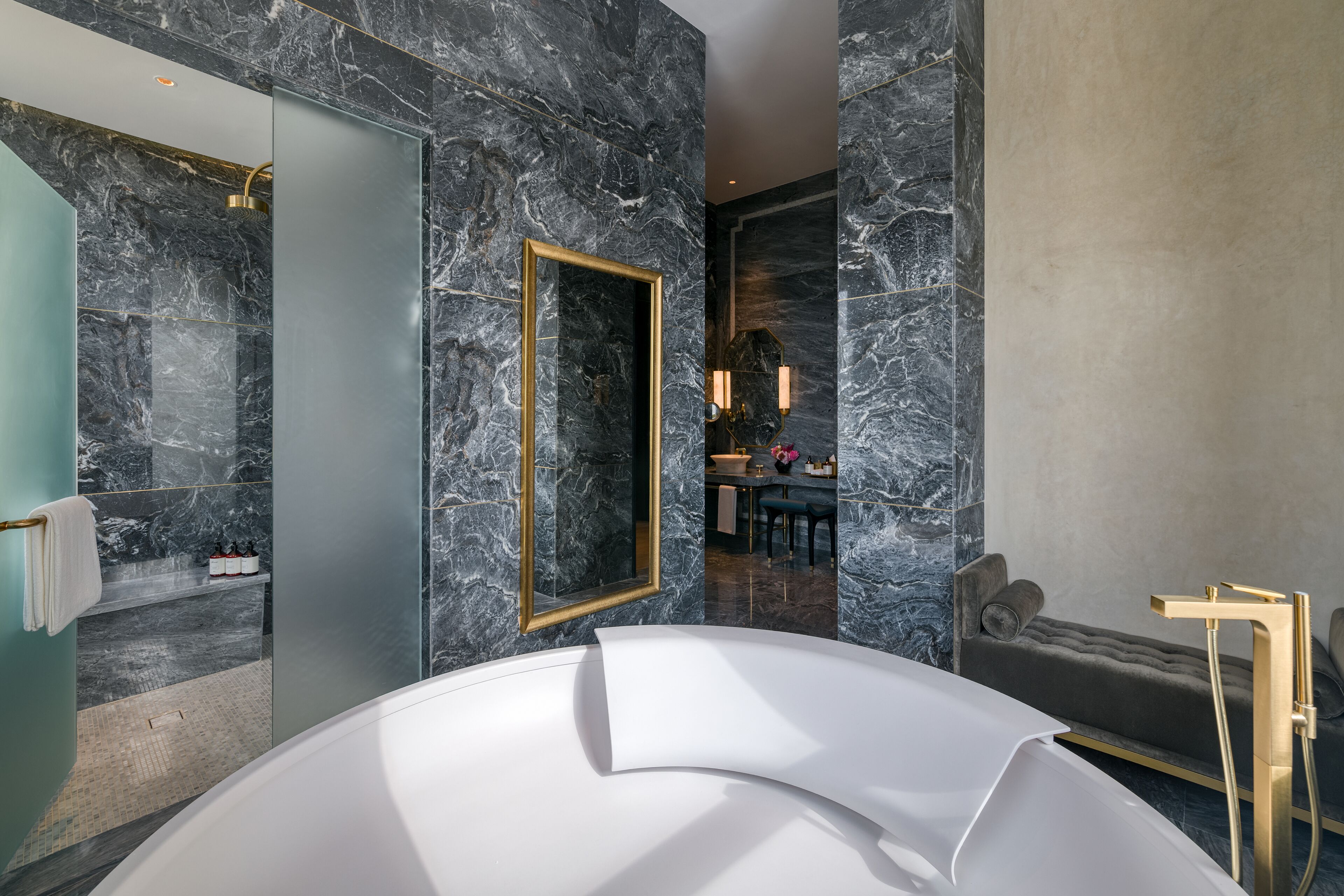 A modern bathroom with a large bathtub, marble walls, a gold-framed mirror, and a separate seating area with a cushioned bench.