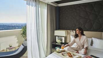A hotel room with a bed, a bedside table with a lamp, a tray with breakfast items, a window with curtains, and a view of the cityscape.