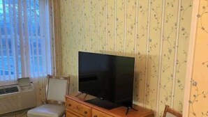 Een kamer met een televisie op een houten dressoir, twee stoelen, een raam met gordijnen en bloemenbehang.