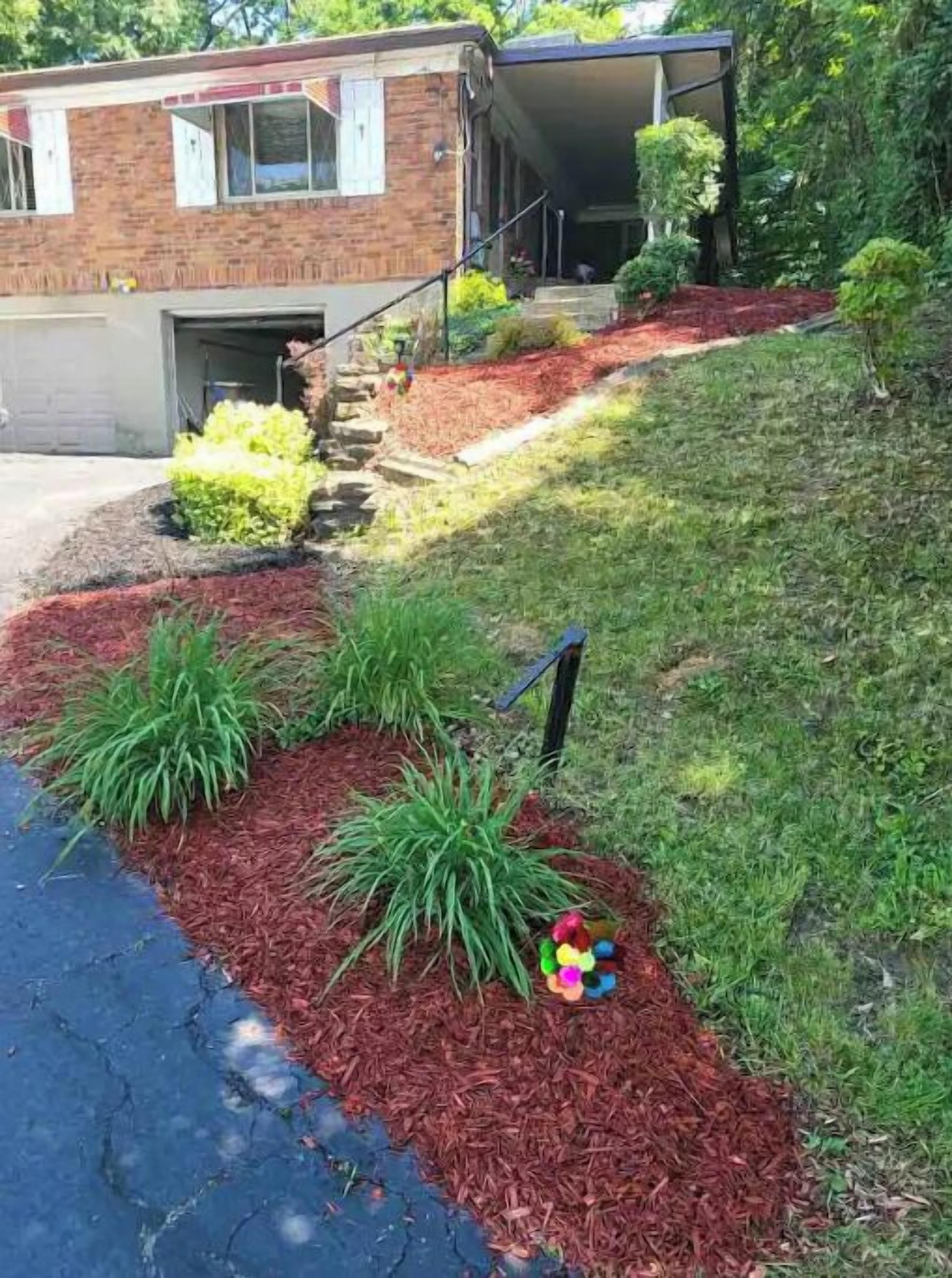 Un quartiere residenziale con una casa in mattoni, vialetto e giardino curato con pacciamatura e piante.