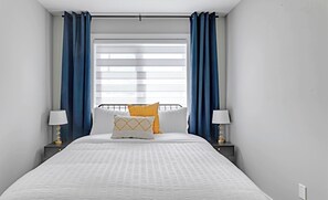A neatly made bed with white bedding and two pillows, flanked by nightstands with lamps, in a room with a window and blue curtains.