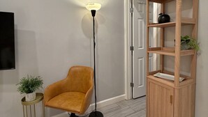 A room with a wooden floor, a tall floor lamp, a leather chair, a small side table with a plant, and a wooden shelving unit.