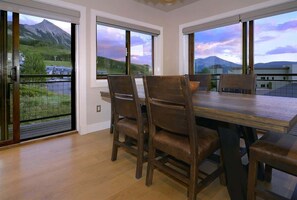 Un comedor con mesa y sillas de madera, ventanas amplias con vista a la montaña y un balcón con barandilla.