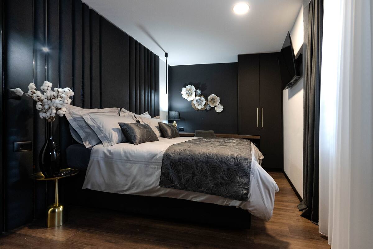 Une chambre moderne avec un lit, un lambris en bois foncé, une table de chevet, un vase de fleurs et un téléviseur fixé au mur.