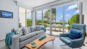 Una sala moderna con un sofá gris, un sillón azul, una mesa de centro de madera y puertas de cristal grandes que dan a una zona de piscina.
