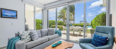 Uma sala de estar moderna com um sofá cinzento, uma poltrona azul, uma mesa de centro de madeira e grandes portas de vidro que dão para uma área de piscina.