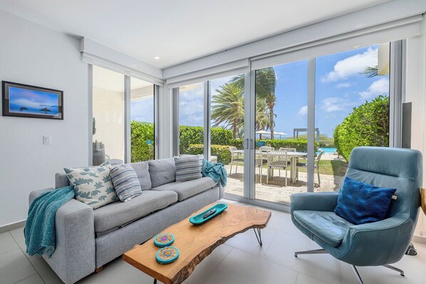 A modern living room with a grey sofa, a blue armchair, a wooden coffee table, and large glass doors leading to a pool area.