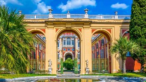 Una entrada imponente con un arco central, flanqueado por estatuas y palmeras, que conduce a un edificio de fachada roja bajo un cielo azul.