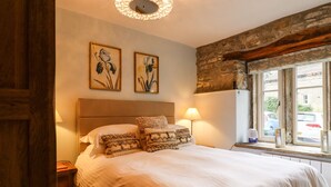 A bedroom with a large bed, two wall-mounted paintings, a wooden headboard, a bedside lamp, and a window with a view of the street. - Squint Cottage (Skipton)