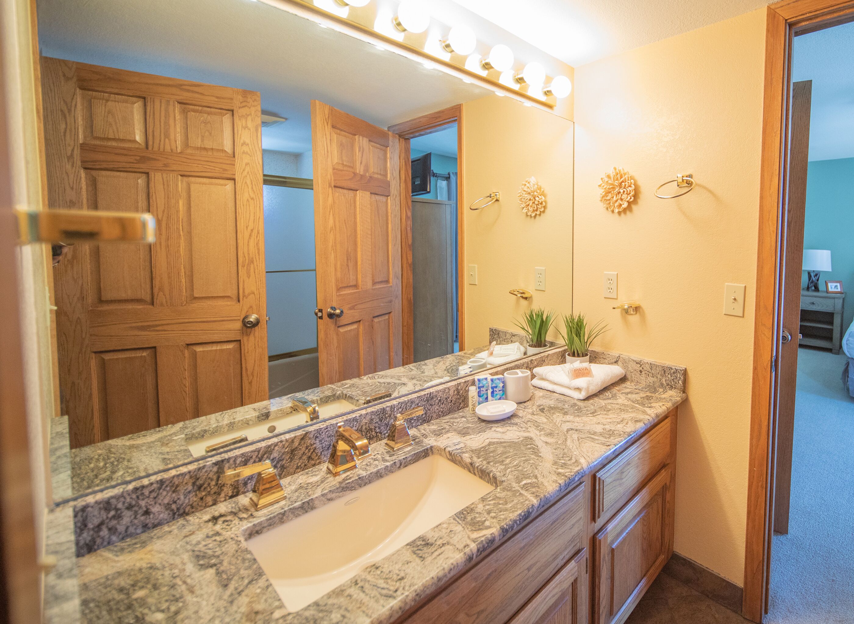 Une salle de bain avec un lavabo en marbre, des meubles en bois, un miroir de coiffeuse et un espace douche visible à travers une porte ouverte.
