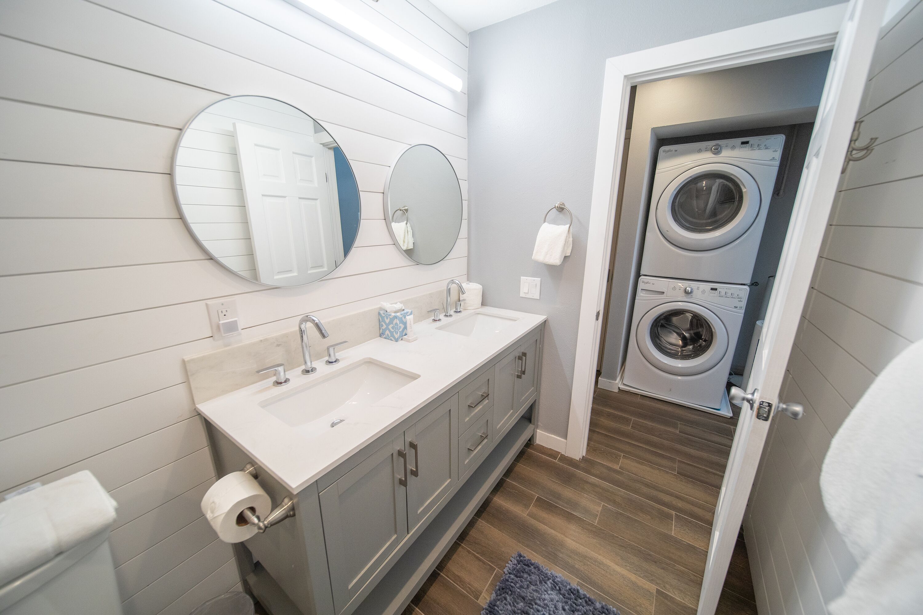 A modern bathroom with a double sink vanity, two mirrors, a toilet paper holder, and a washing machine in the adjacent room.