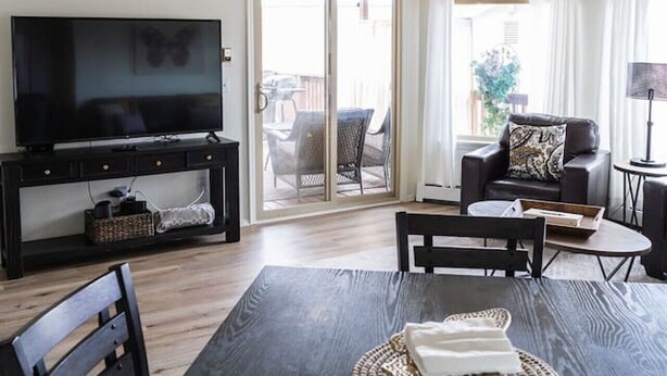 A living room with a black TV stand, a dining table with chairs, and a sofa with a patterned throw pillow. There is a chandelier with multiple white lampshades. - The Hillview Hangout (Grand Junction)