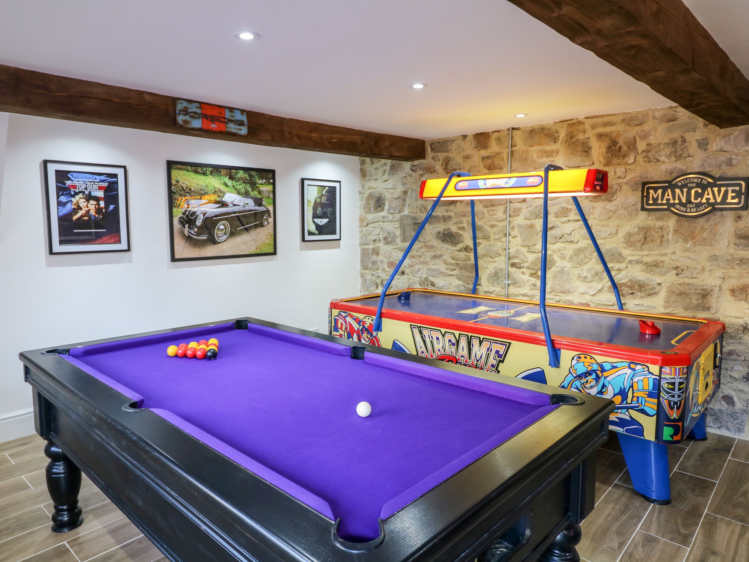 A pool table with a purple felt surface, a billiards cue, and billiard balls. There are framed pictures on the wall, a wooden ceiling, and a stone wall.