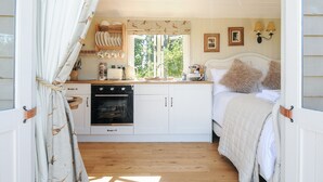 A cozy bedroom with a bed, pillows, and a bedside table. There is a window with curtains, a sink, and a small kitchen area with a stove and cabinets.