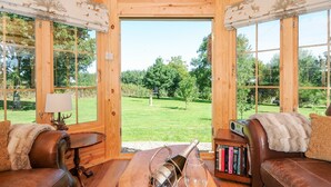 A cozy room with a wooden ceiling, large windows, a leather sofa, a wooden table with a bottle and glasses, and a bookshelf.