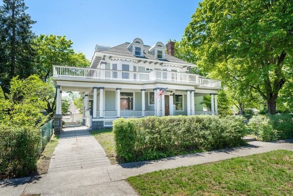 A two-story colonial-style house with a front porch, a small balcony, and a flag. - Historic Family Home with 8 BDRMS, 8 BATHS & 3 Kitchens! (Spokane)