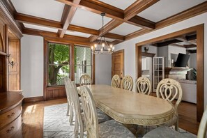 A dining room with a large wooden table and six chairs, a chandelier, and a view of the outdoors through large windows. - Historic Family Home with 8 BDRMS, 8 BATHS & 3 Kitchens! (Spokane)