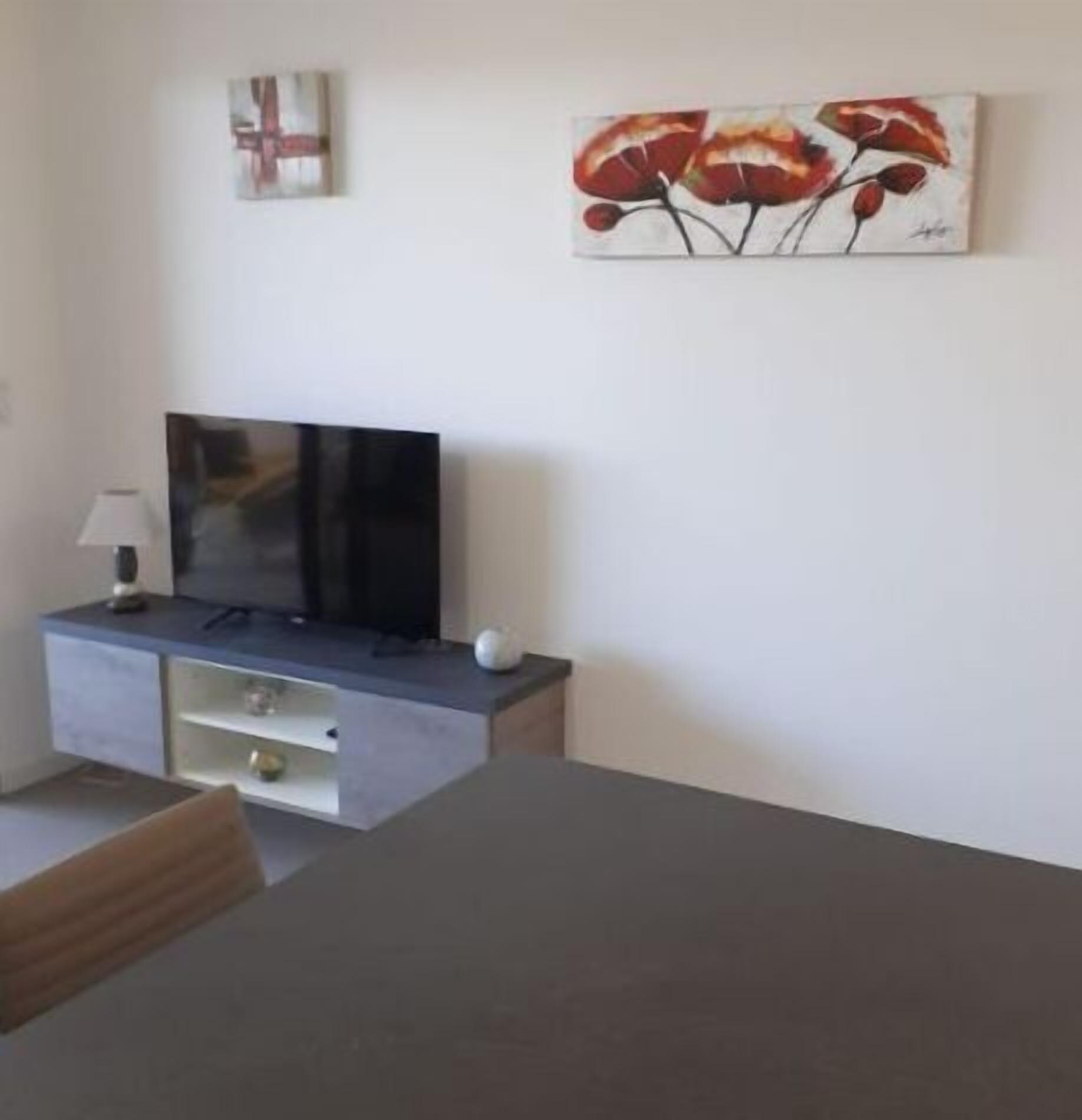 Ein modernes Wohnzimmer mit Fernseher auf grauem Schrank, einem Esstisch mit Stühlen und einem Gemälde von roten Blumen an der Wand.