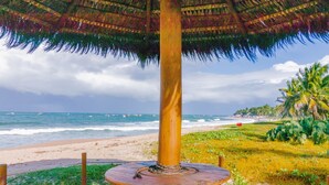Une scène de plage avec une table en bois placée sous un parasol en chaume, un rivage sablonneux et des palmiers.