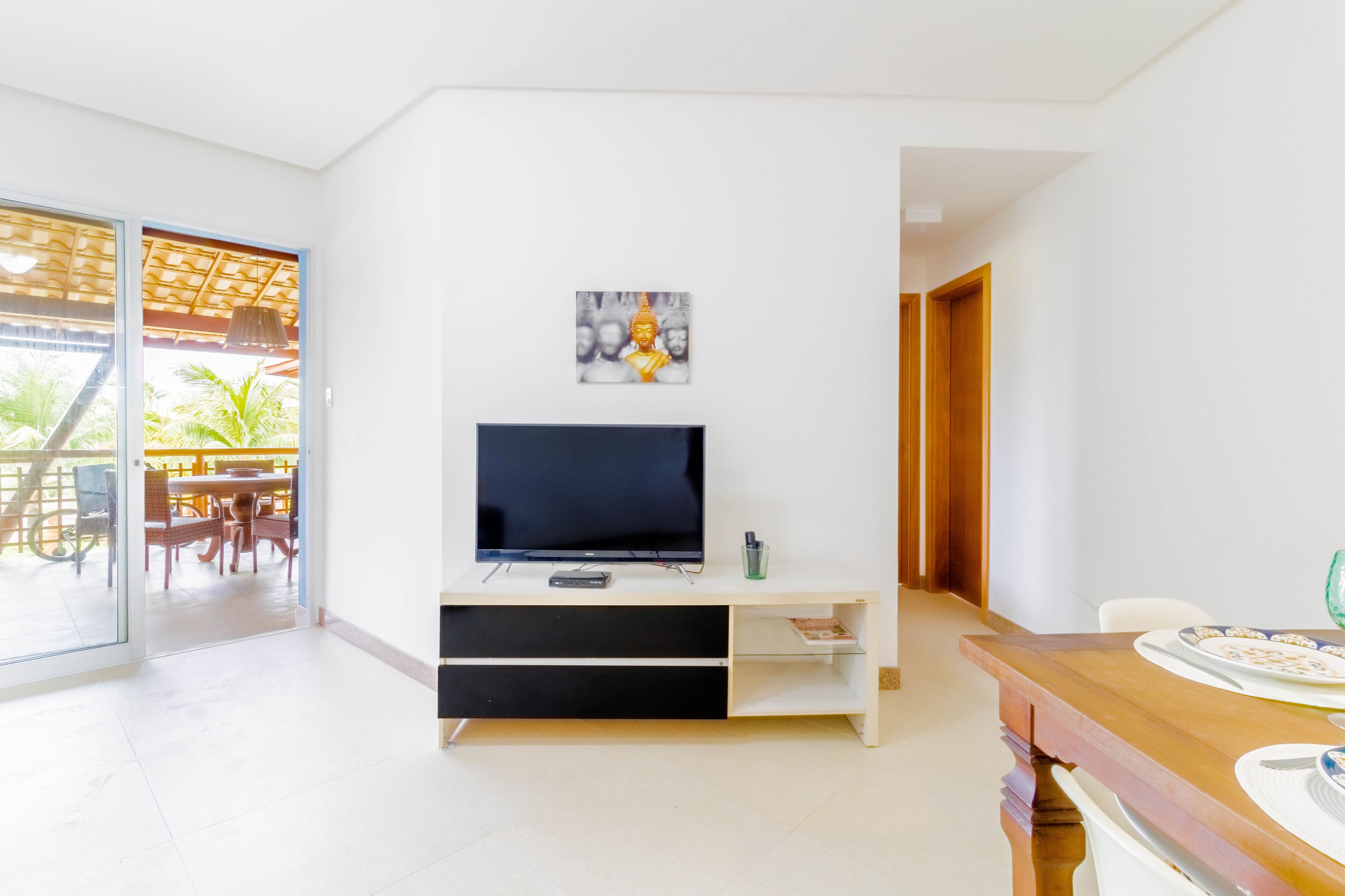Un salon moderne avec un téléviseur à écran plat posé sur un meuble de télévision blanc, un coin repas avec une table et des chaises en bois, et une porte-fenêtre coulissante donnant sur une terrasse extérieure.