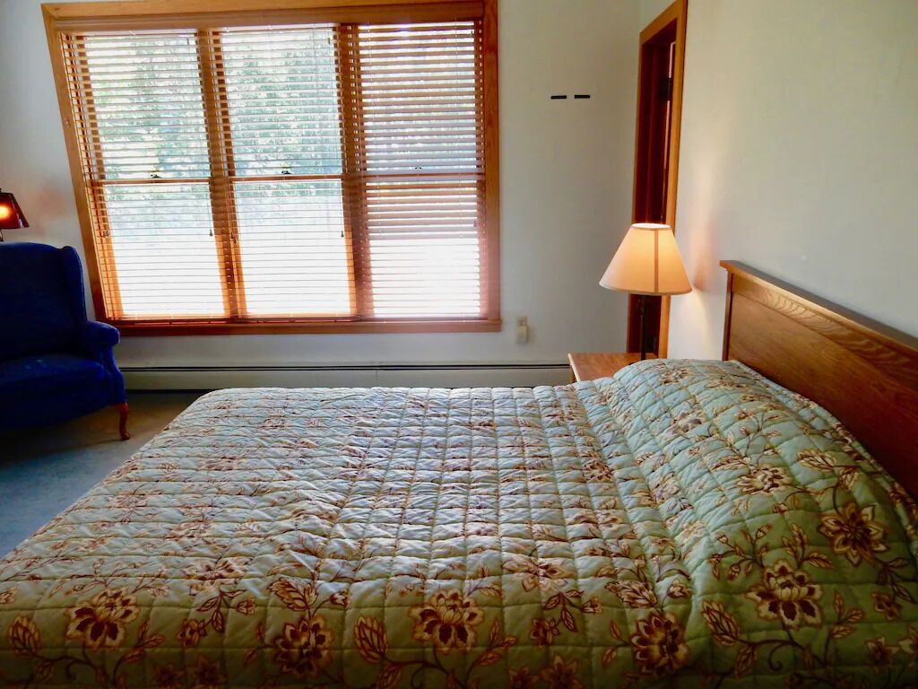Ein Schlafzimmer mit einem großen Bett, einem Fenster mit Jalousien, einer Lampe und einem blauen Sessel.