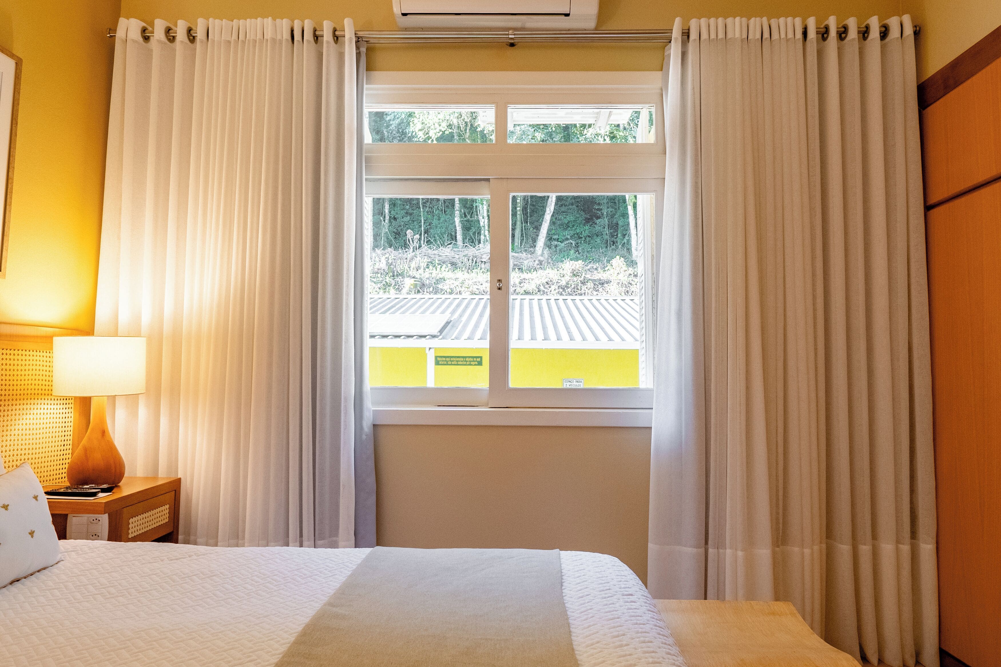 Una habitación de hotel con cama, mesita de noche, lámpara y ventana con cortinas.