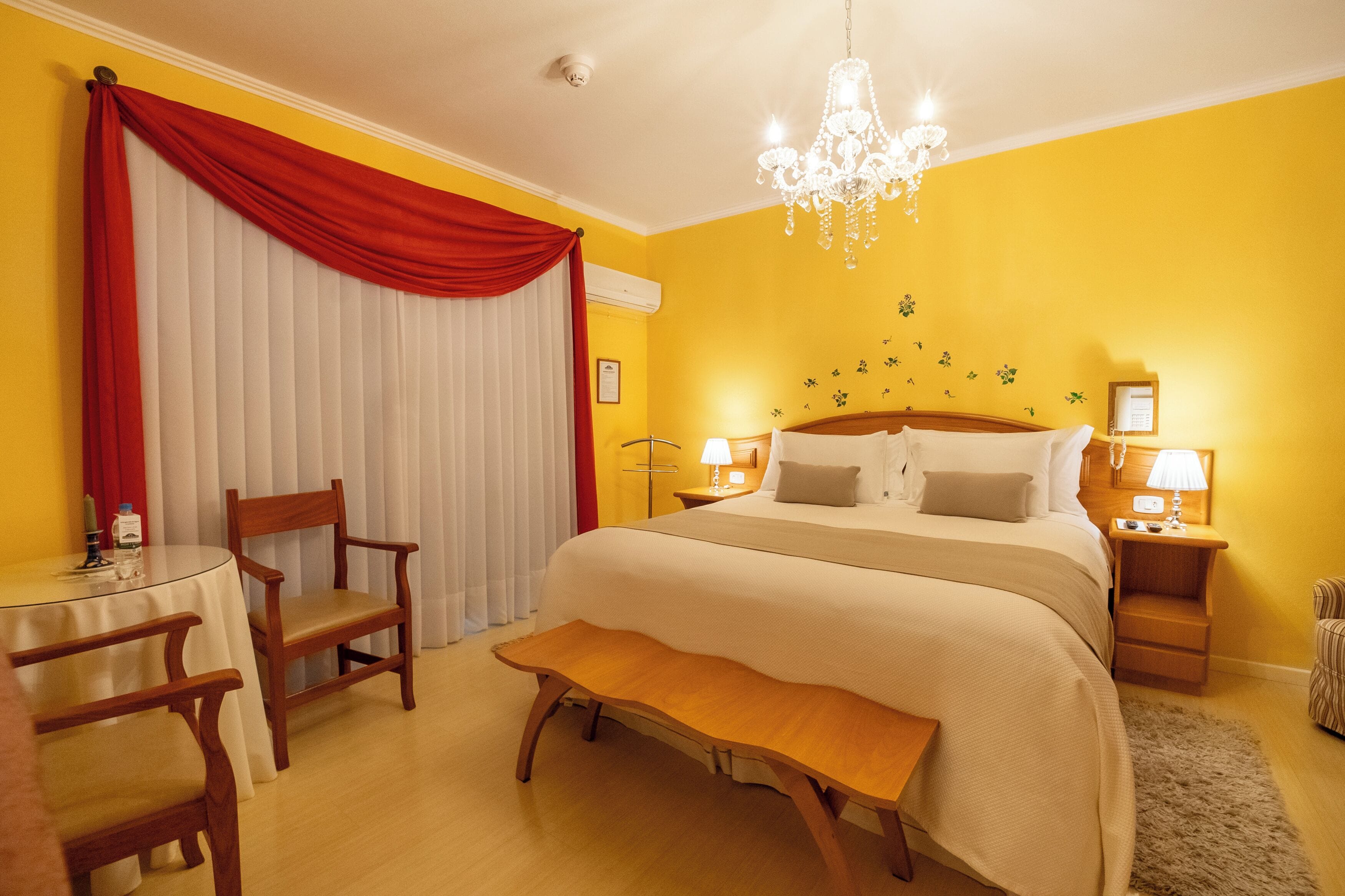 Una habitación de hotel con una cama grande, un banco de madera, una mesa redonda con sillas y una araña.