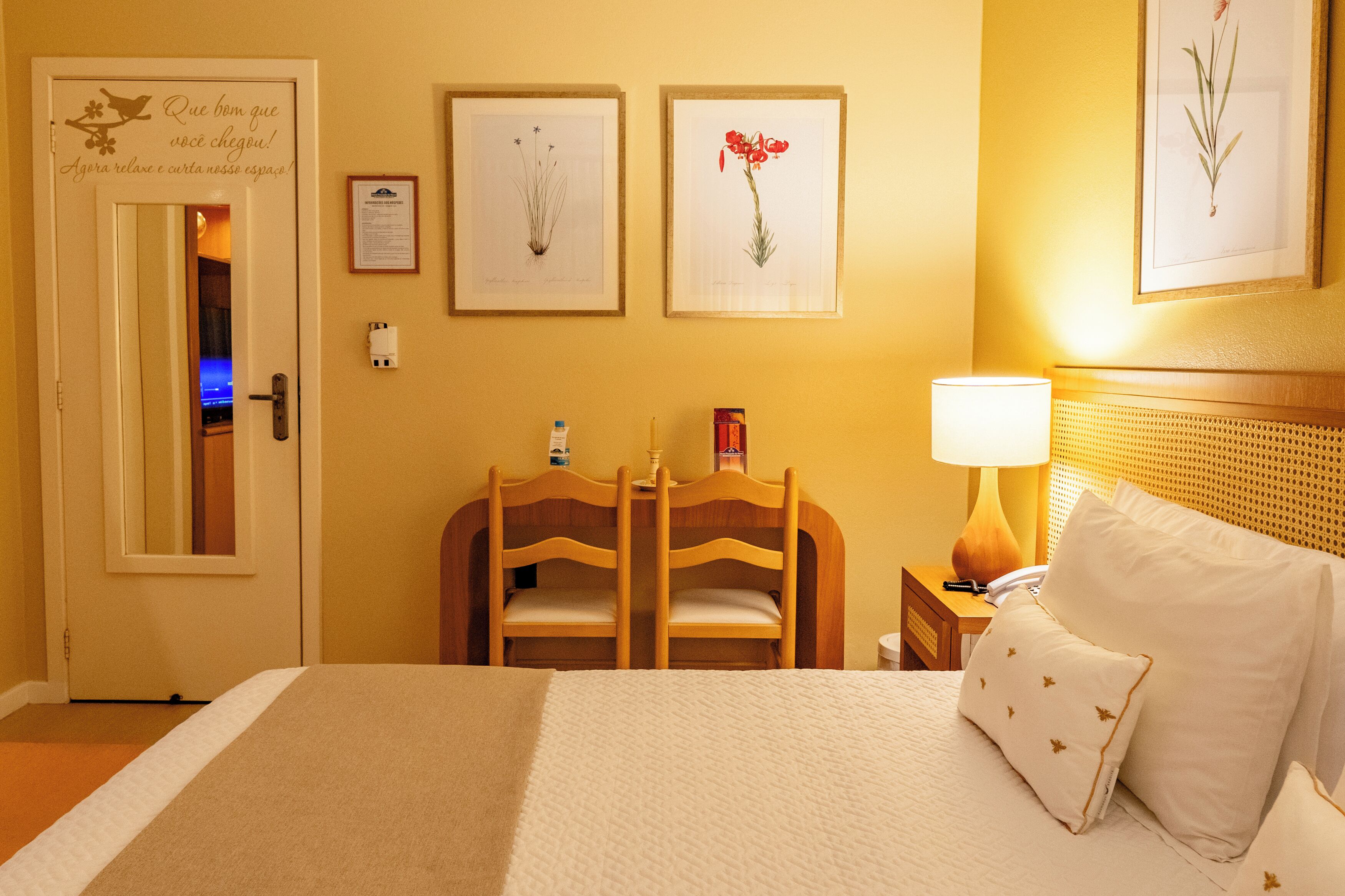 A hotel room with a bed, a wooden chair, a nightstand with a lamp, and framed botanical prints on the wall.