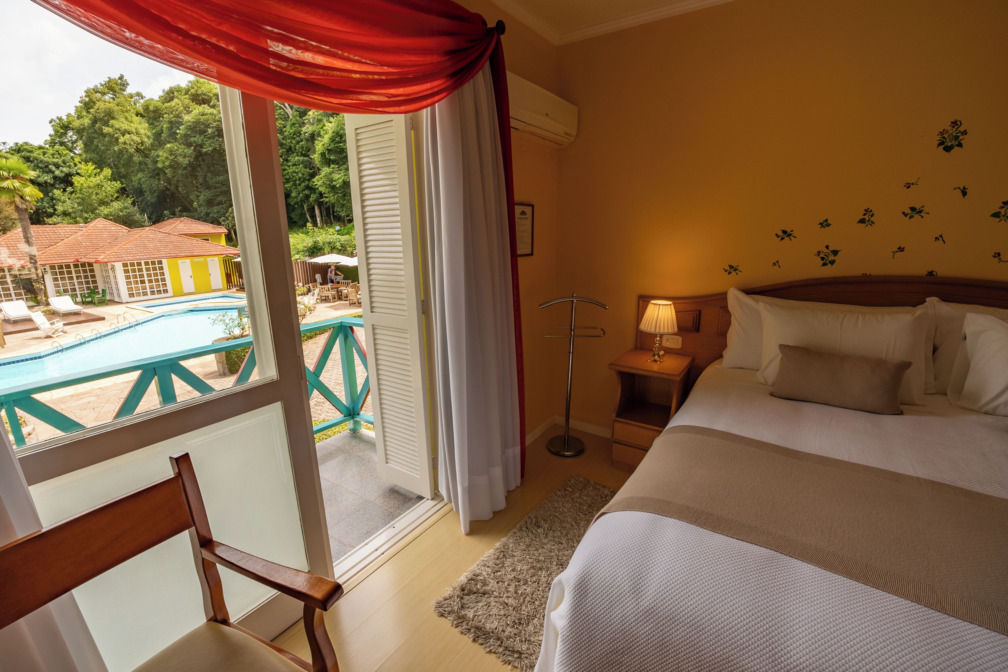 Hotelkamer met een bed, nachtkastje, lamp, uitzicht op een zwembad en een balkon met een rood gordijn.