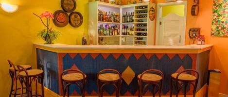 Zona de bar con barra, estantes llenos de botellas y cinco taburetes de mimbre.