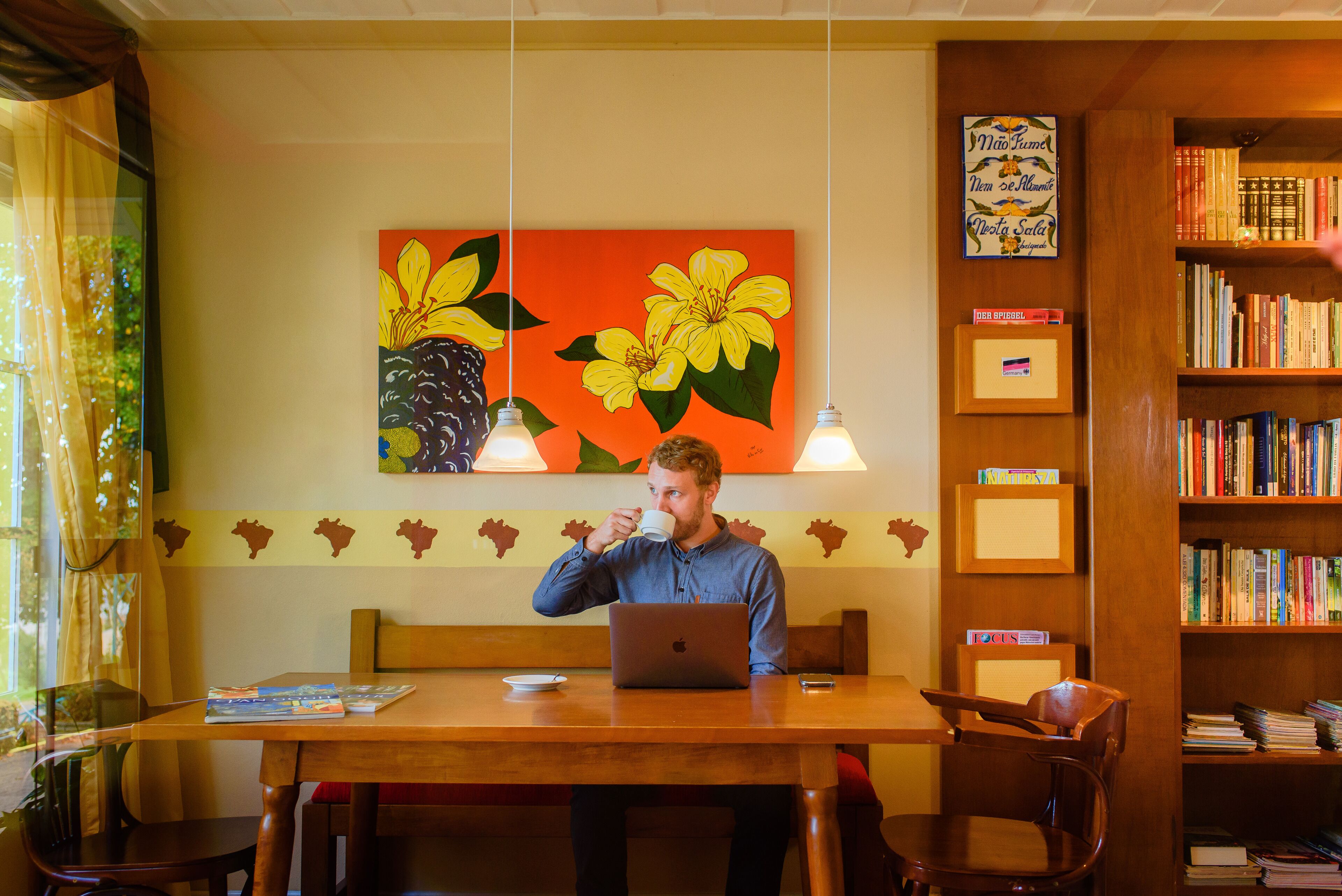 A person sitting at a wooden table with a laptop, a cup, and a book. There is a bookshelf with books and a framed picture on the wall.