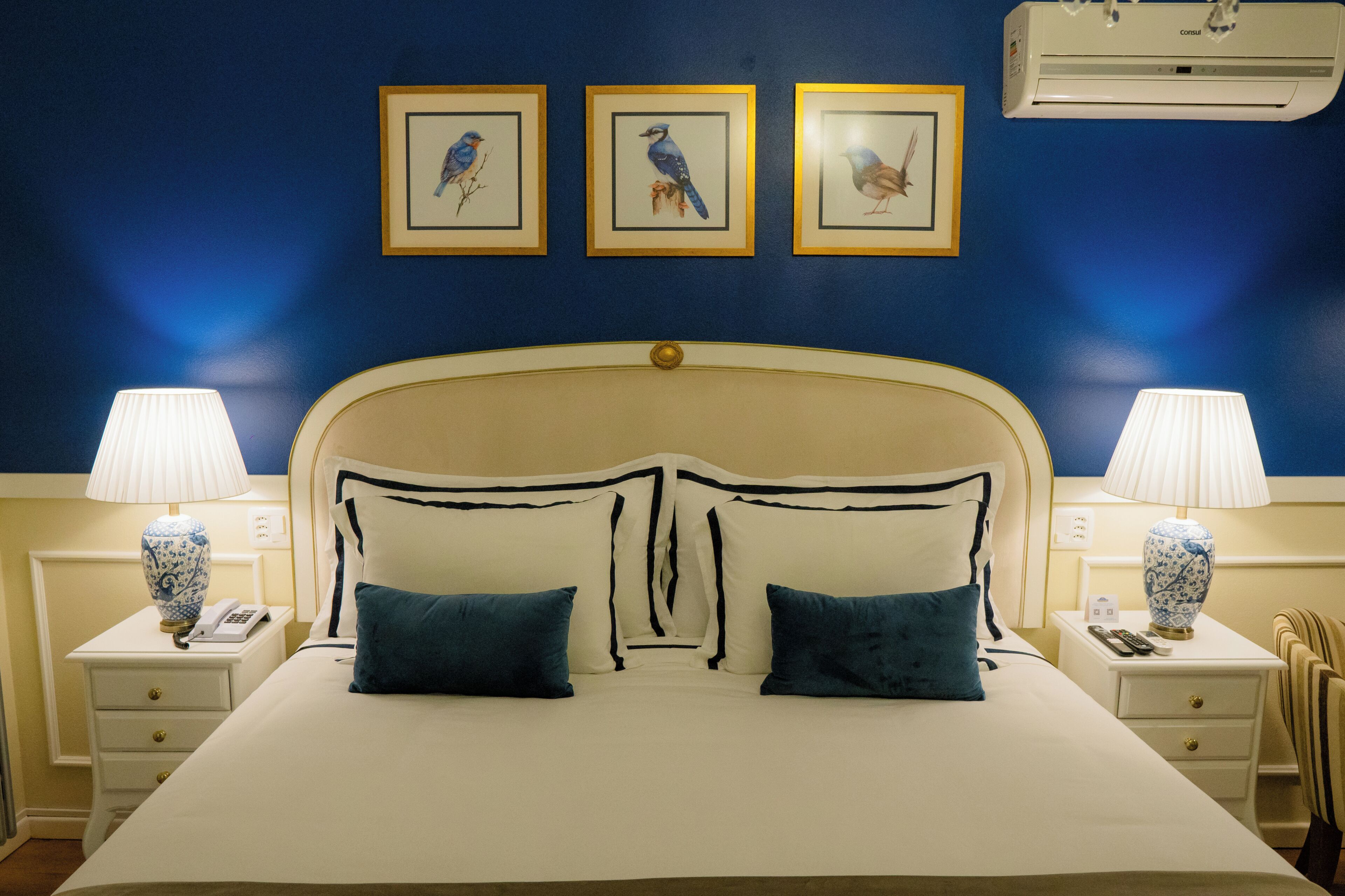 Una cama bien tendida con cabecera, dos mesitas de noche con lámparas, y obras de arte enmarcadas en la pared.